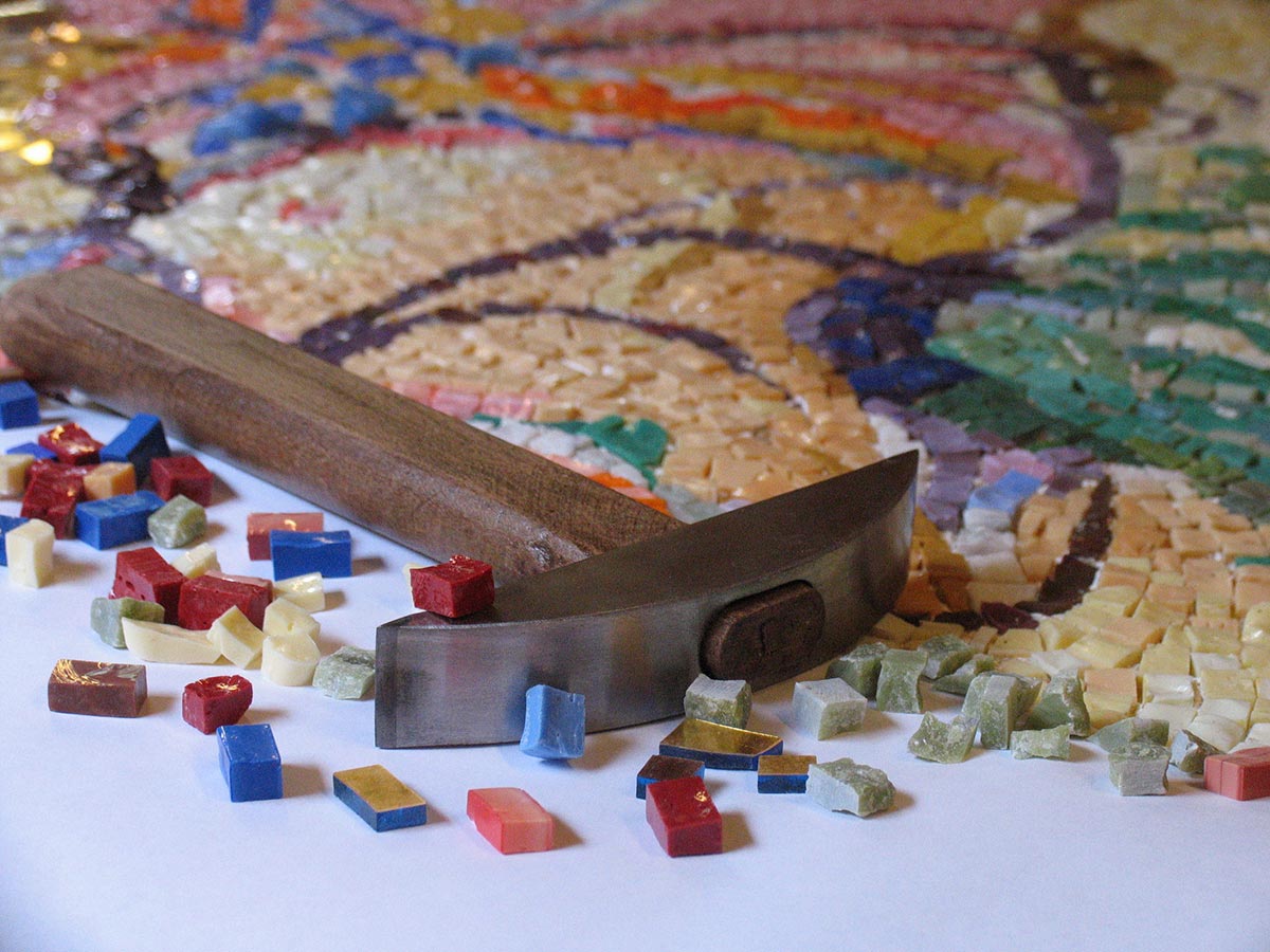 Primo piano di una martellina per mosaico artistico appoggiata su un mosaico n costruzione a smalto colorato.